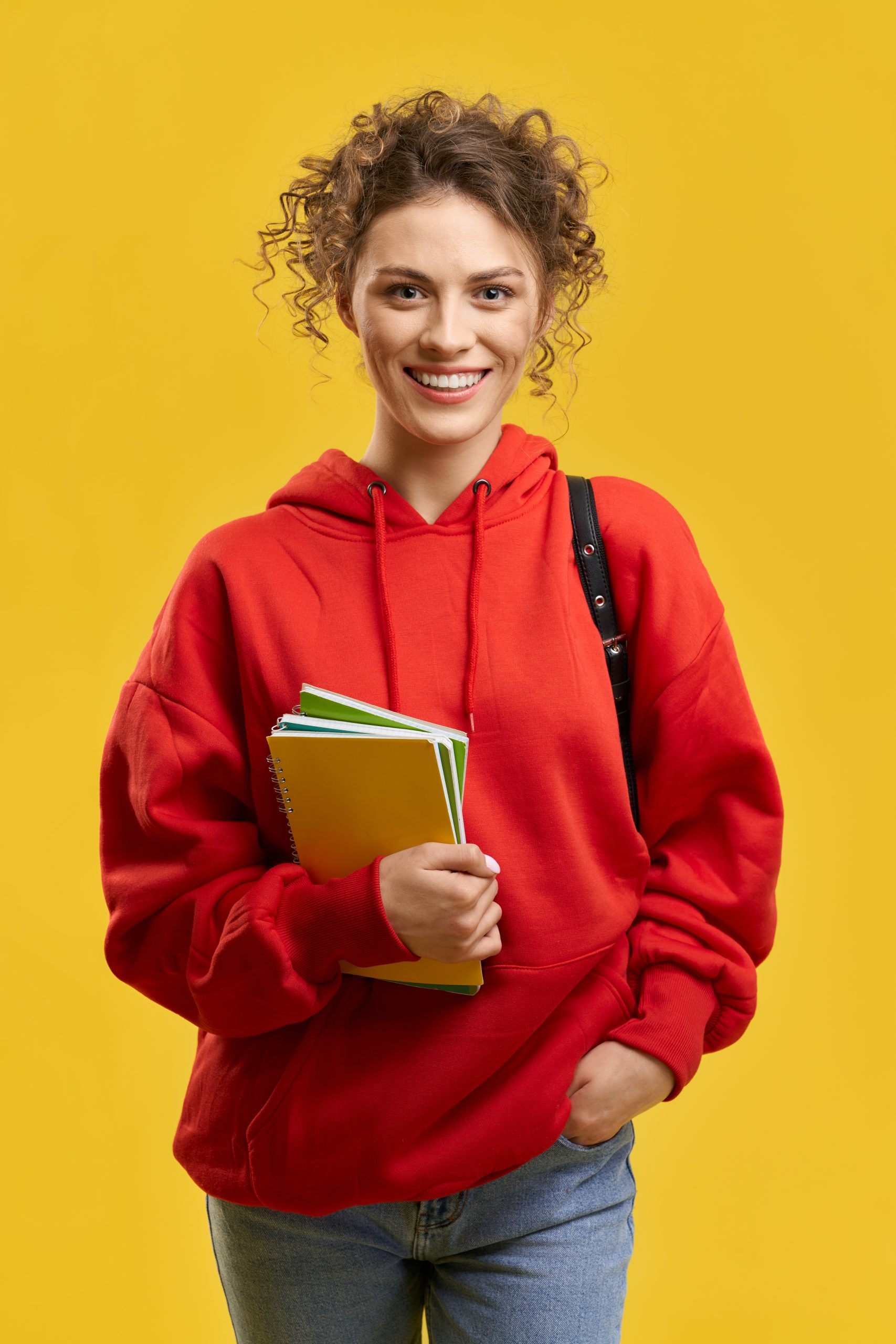 Portrait of girl with curly hair standing, smiling. Student with rucksack and curly hair looking at camera, hlding hand in pocket, wearing red khudi and jeans, Concept of youth.